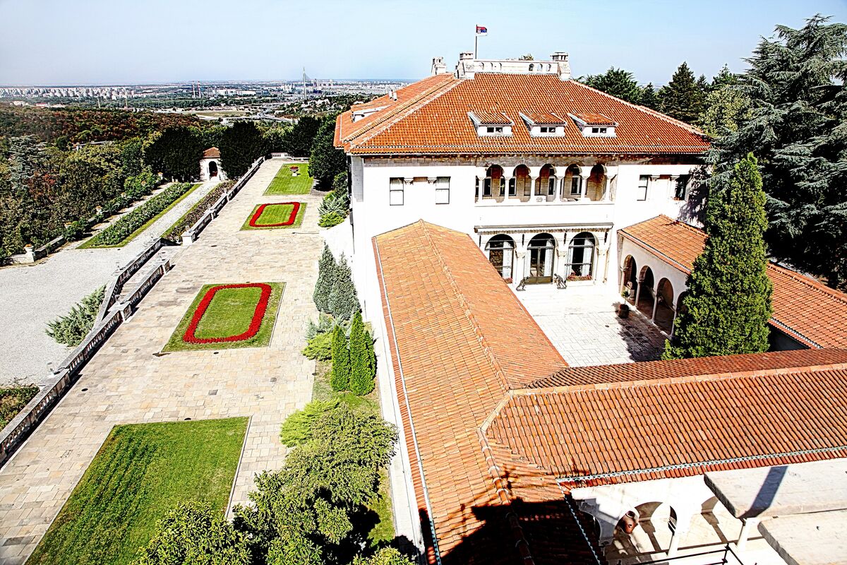 The Royal Palace in Dedinje seen from above, with red tile roofs, an arcaded courtyard and terraced gardens, and New Belgrade on the horizon