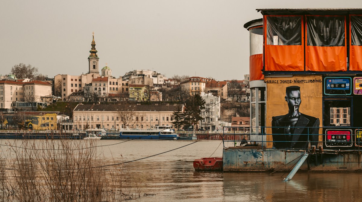 A floating club with a painted mural moored on the Sava, with the old town and the Cathedral bell tower behind