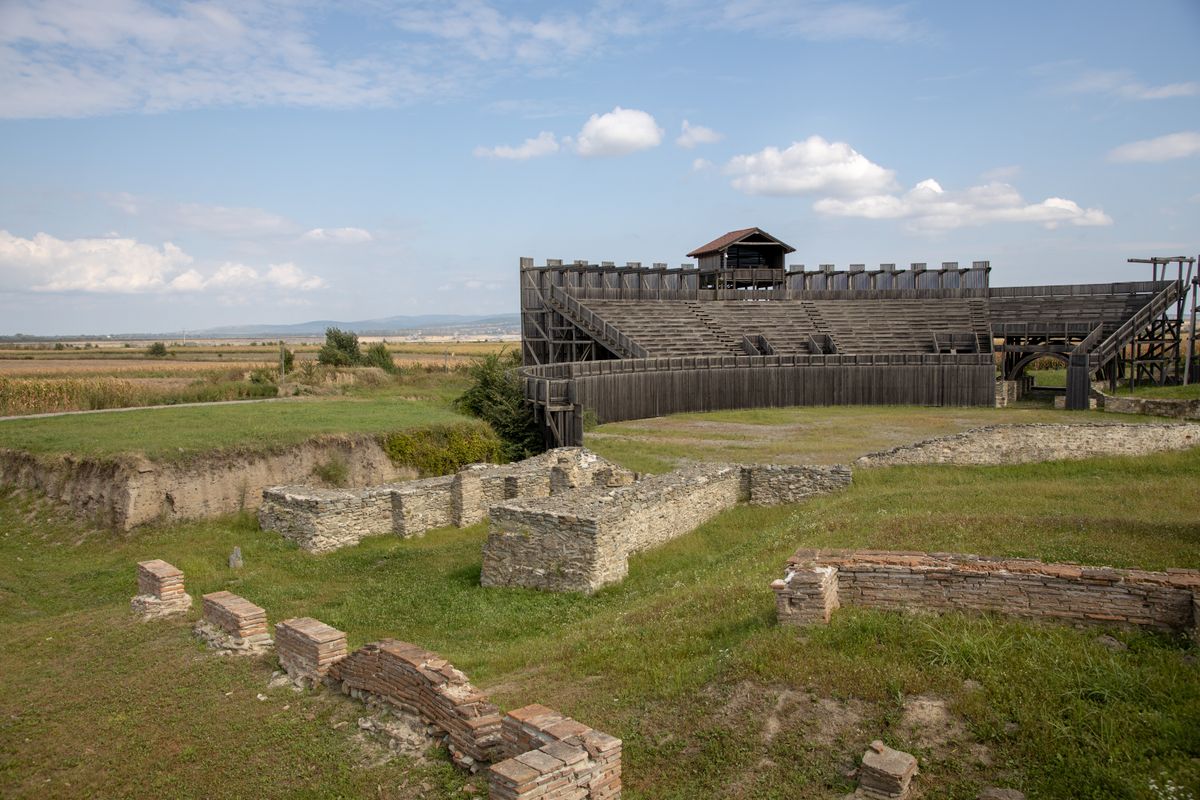 Excavated Roman walls in the grass at Viminacium, with a reconstructed wooden amphitheater behind them