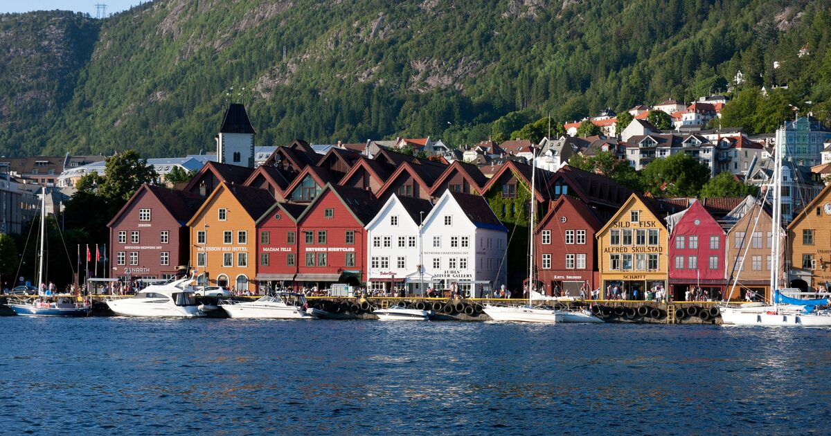 The colorful wooden Hanseatic houses of Bryggen along Bergen's harbor, with boats in front and a forested mountain behind