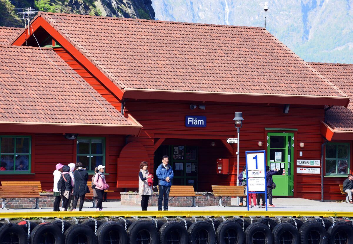 The red wooden railway station of Flåm with travelers on the platform and mountains behind