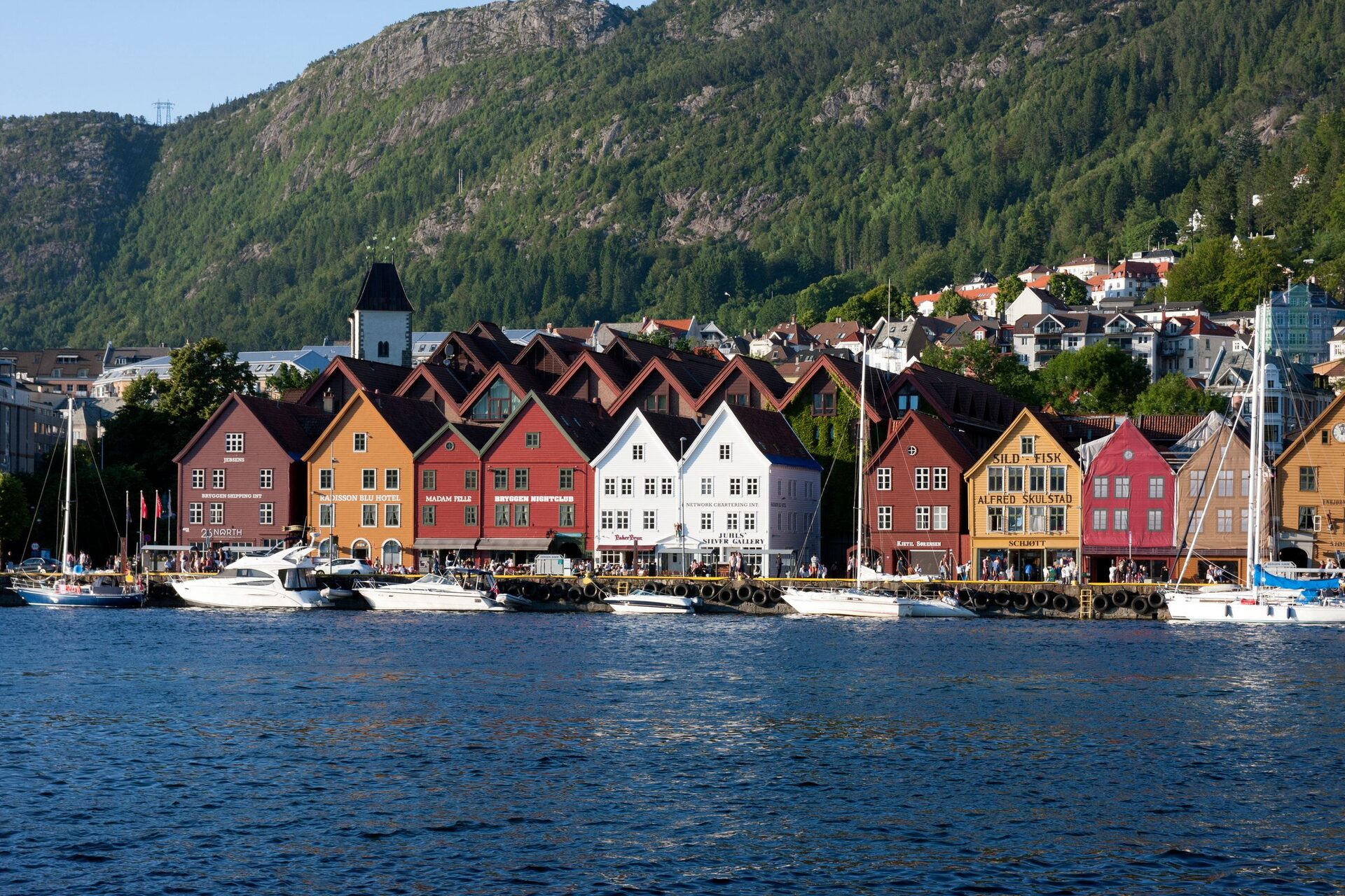 The colorful wooden Hanseatic houses of Bryggen along Bergen's harbor, with boats in front and a forested mountain behind