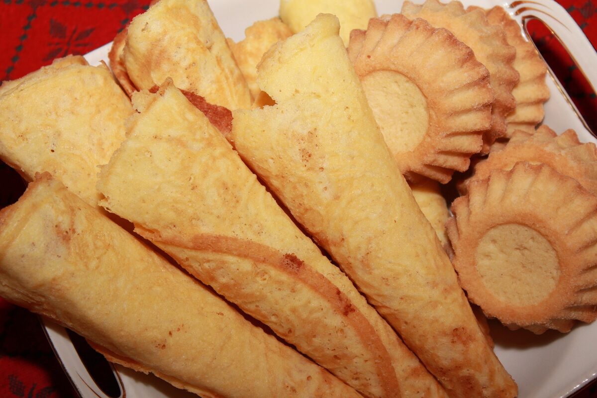 Crisp golden krumkake cones piled on a plate with small fluted sand cookies