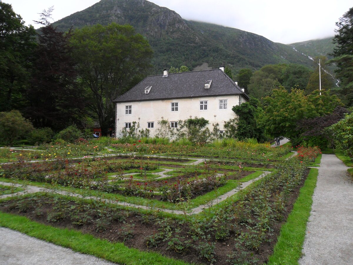 The white manor house of Baroniet Rosendal behind a formal rose garden, with green mountains behind