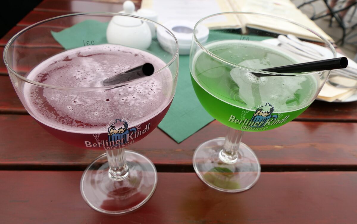 Two glasses of Berliner Weisse mit Schuss, one with red raspberry syrup and one with green woodruff syrup
