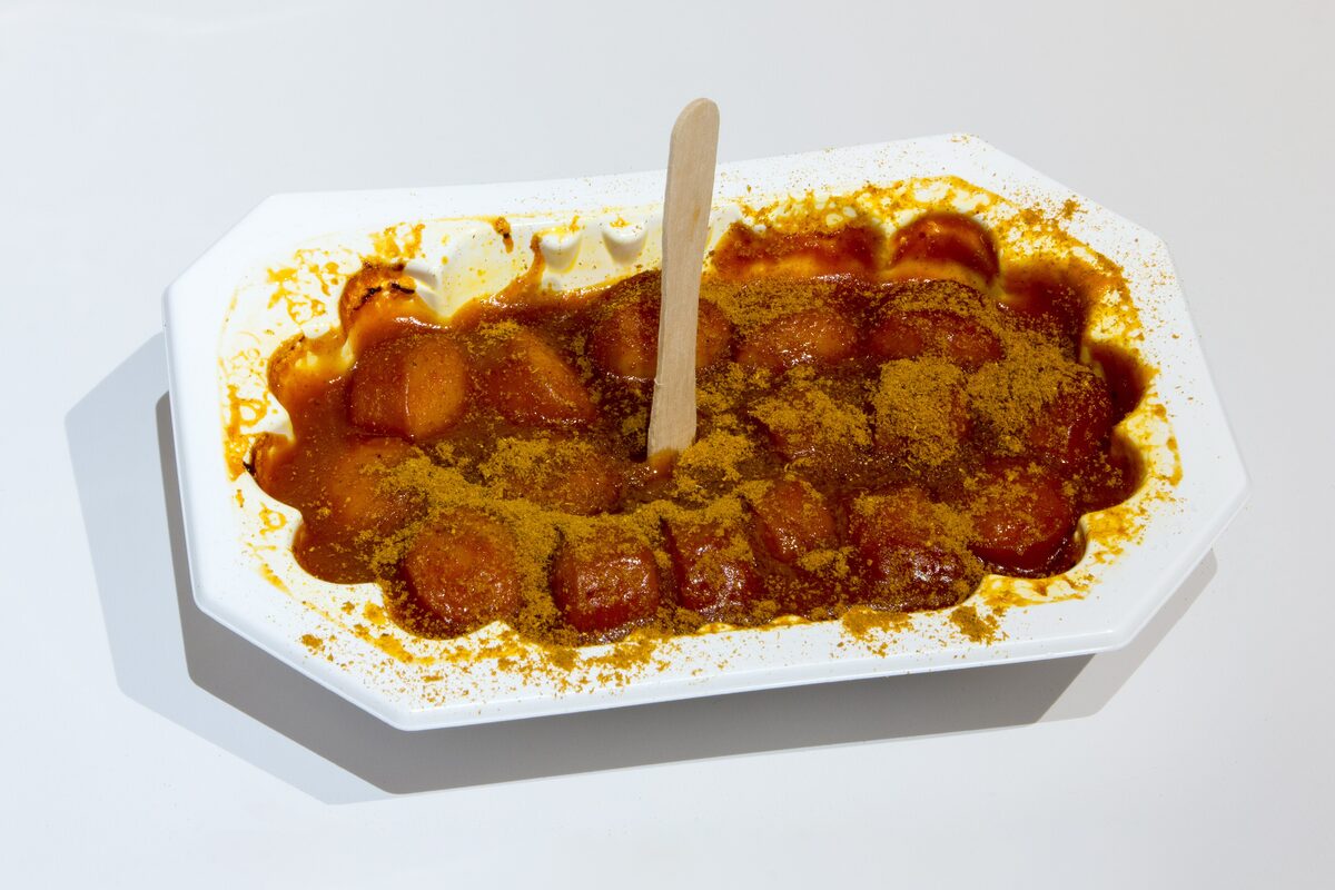 A paper tray of sliced currywurst under ketchup sauce and curry powder, with a wooden fork
