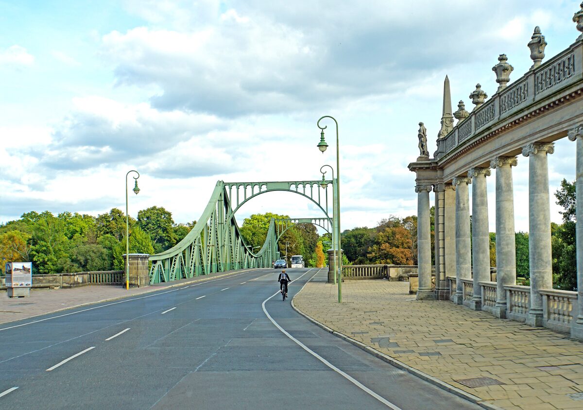 The green steel Glienicke Bridge seen from the Potsdam side, with a colonnade on the right and a cyclist on the road