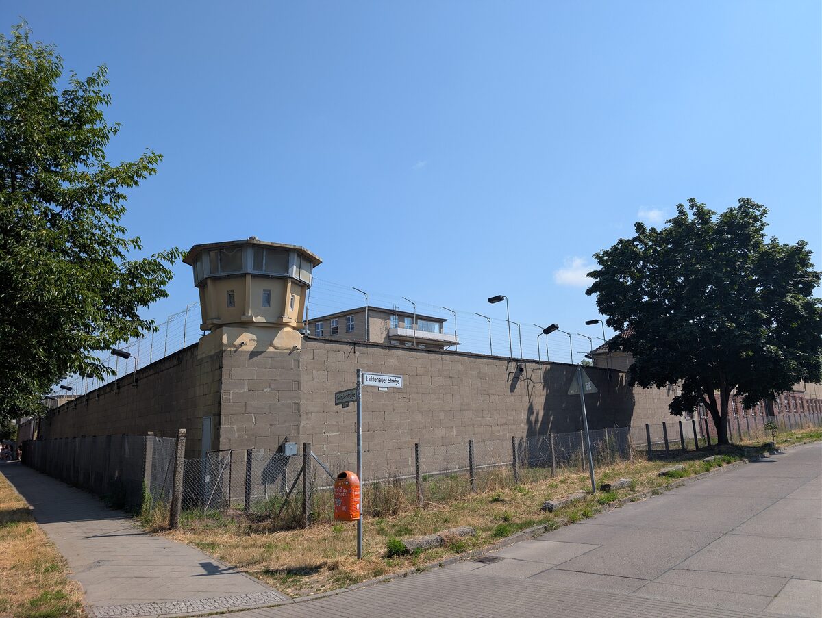 The high concrete wall and guard tower of the former Stasi prison at Hohenschönhausen, seen along a street corner
