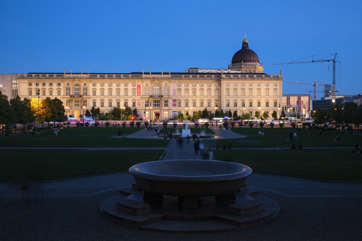 The long facade and dome of the Humboldt Forum, the rebuilt Berlin Palace, lit at dusk beyond a lawn and a stone fountain basin