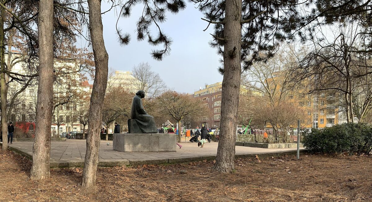 The seated bronze statue of Käthe Kollwitz in Kollwitzplatz among pine and bare trees, with playground and apartment buildings behind