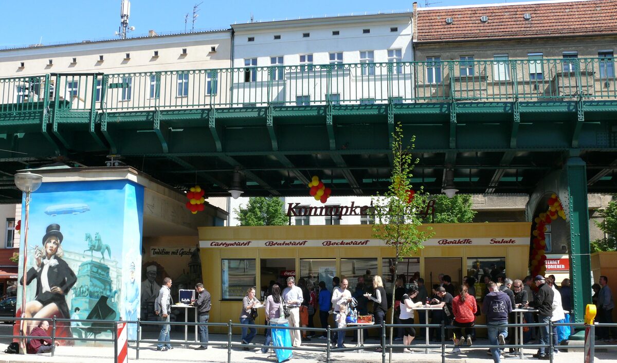 Customers queuing at Konnopke's Imbiss, a yellow snack stand under the green steel viaduct of the U2 line