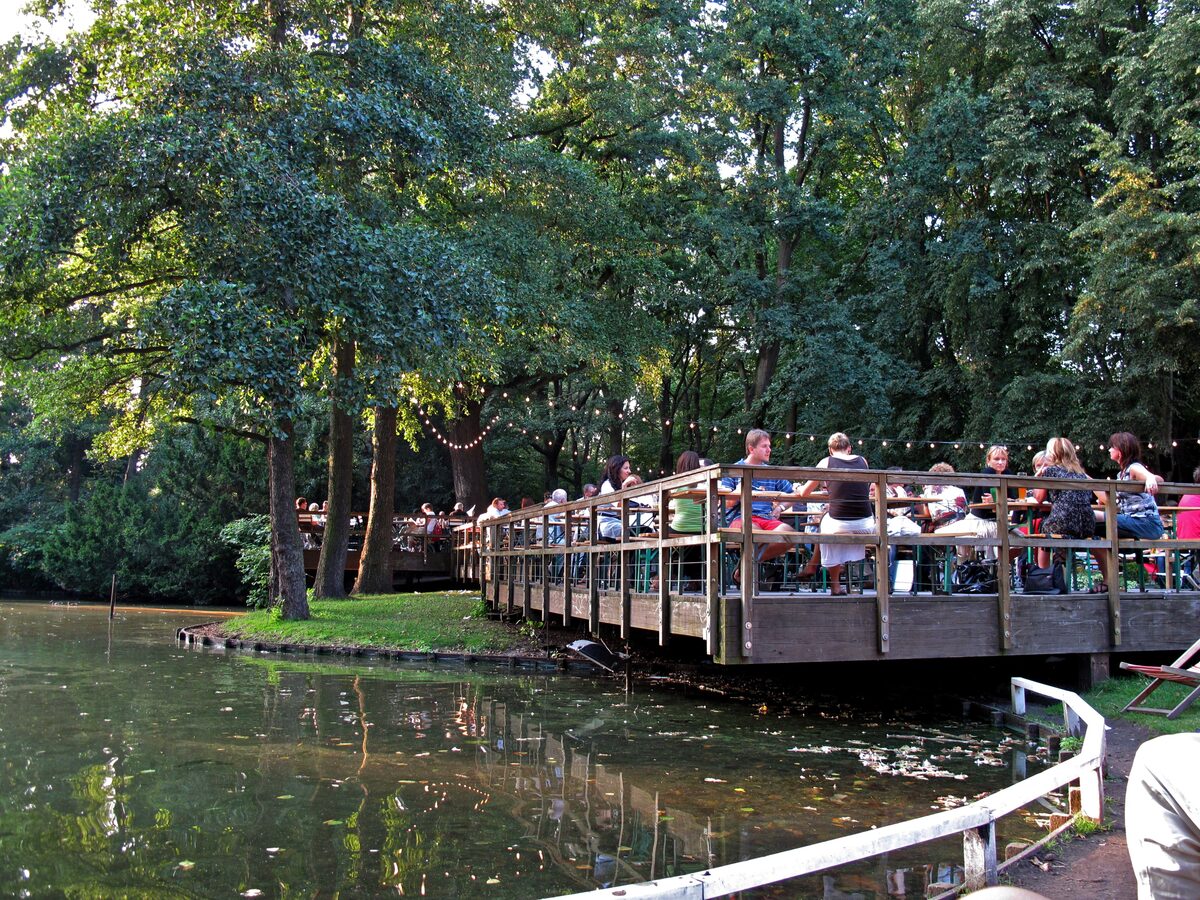 Guests at tables on the wooden terrace of Café am Neuen See, a lakeside beer garden under tall trees in the Tiergarten