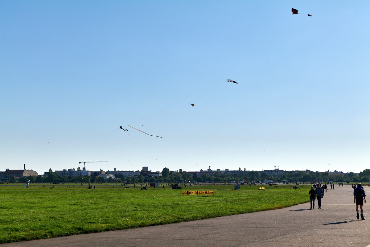 Walkers on a paved runway path across the wide grass of Tempelhofer Feld, with kites flying in the blue sky