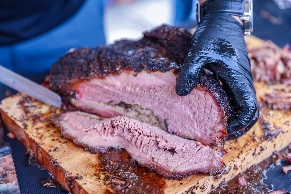 A gloved hand slicing a smoked beef brisket with a dark peppery bark and a pink center on a wooden board