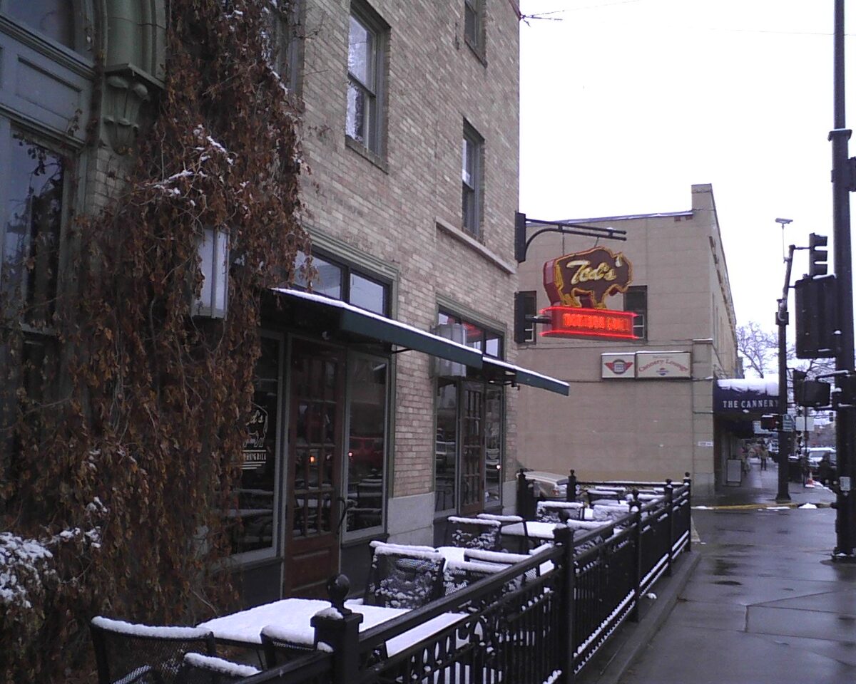 A snow-dusted patio and brick buildings on Main Street in Bozeman, with the neon bison sign of Ted's Montana Grill