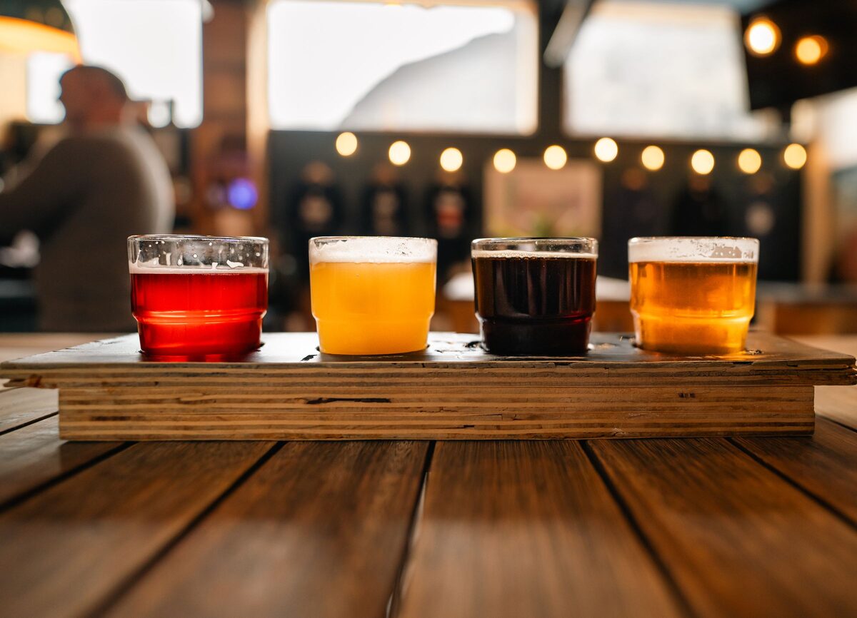 A wooden paddle holding a flight of four craft beers on a table in a taproom, from red amber to pale gold and dark brown