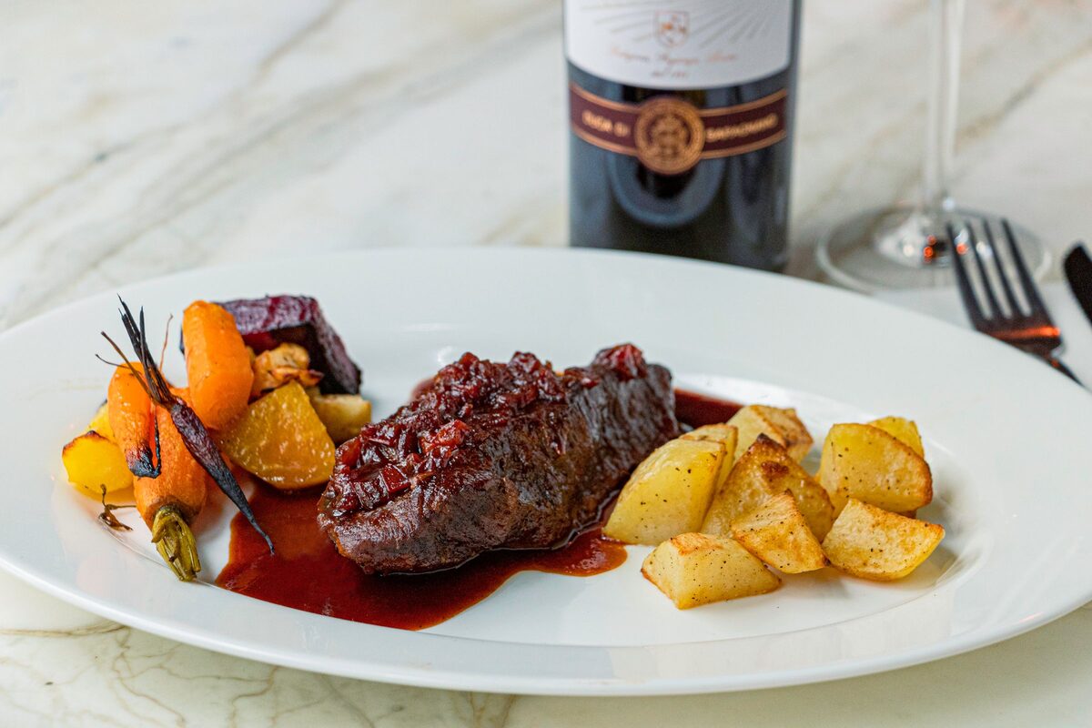 A plated cut of beef in a dark berry sauce with roast carrots and potatoes, a bottle of red wine behind