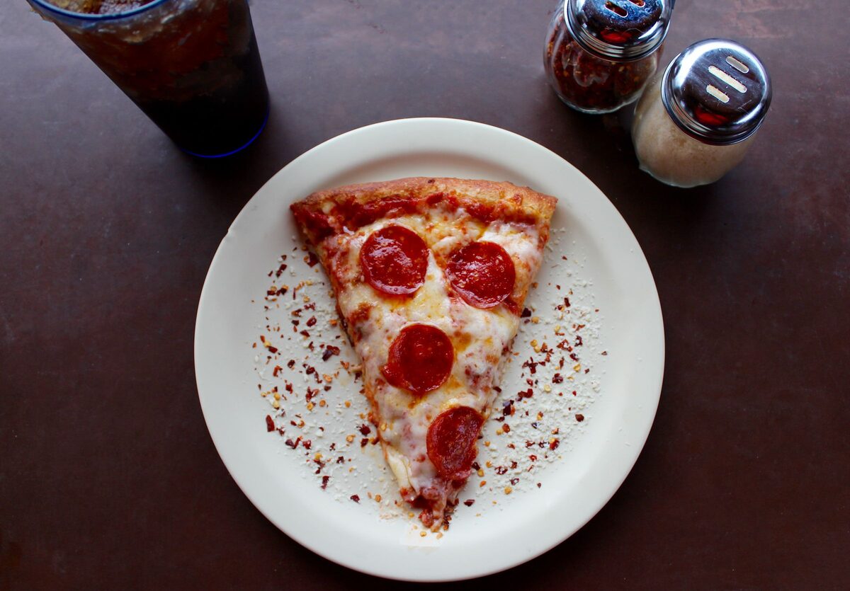 A single slice of pepperoni pizza on a white plate with chili flakes, a soda and shakers on the table