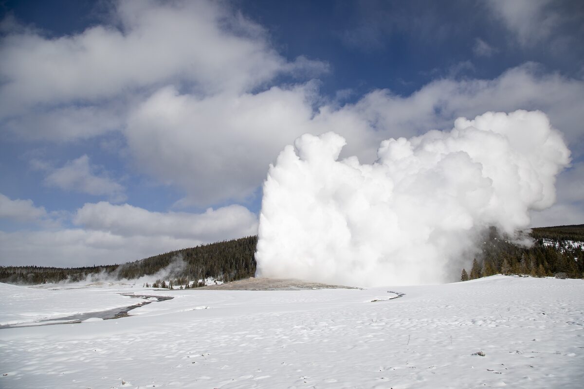Old Faithful erupting over a snowy Upper Geyser Basin under a blue sky with scattered clouds
