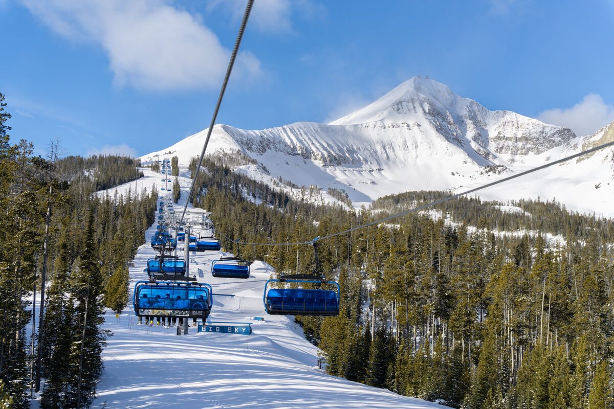Blue bubble chairs climbing a snowy slope through spruce, with Lone Peak white against a blue sky ahead