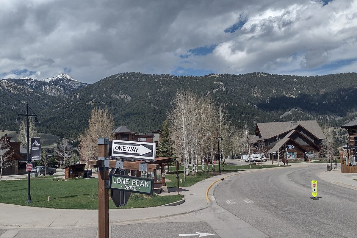 A street sign for Lone Peak Drive in Big Sky Town Center, seen here in late spring without snow, with log-style lodges and forested mountains behind
