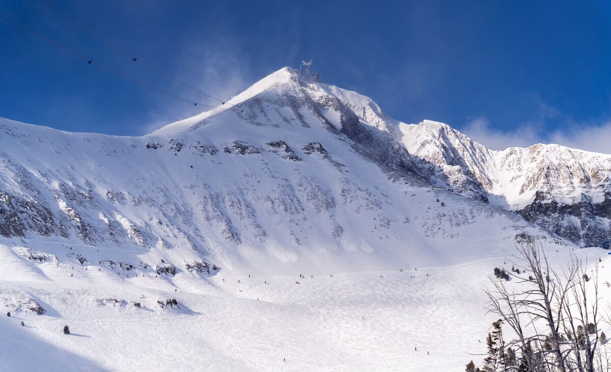 Lone Peak rising above a wide, snowy bowl with a few skiers on the slope, under a deep blue sky