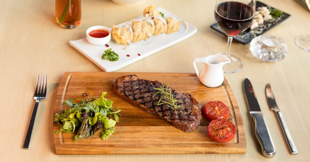 A grilled steak with a rosemary sprig and roasted tomatoes on a wooden board, a glass of red wine and a plate of sushi rolls beside it (generic restaurant table)