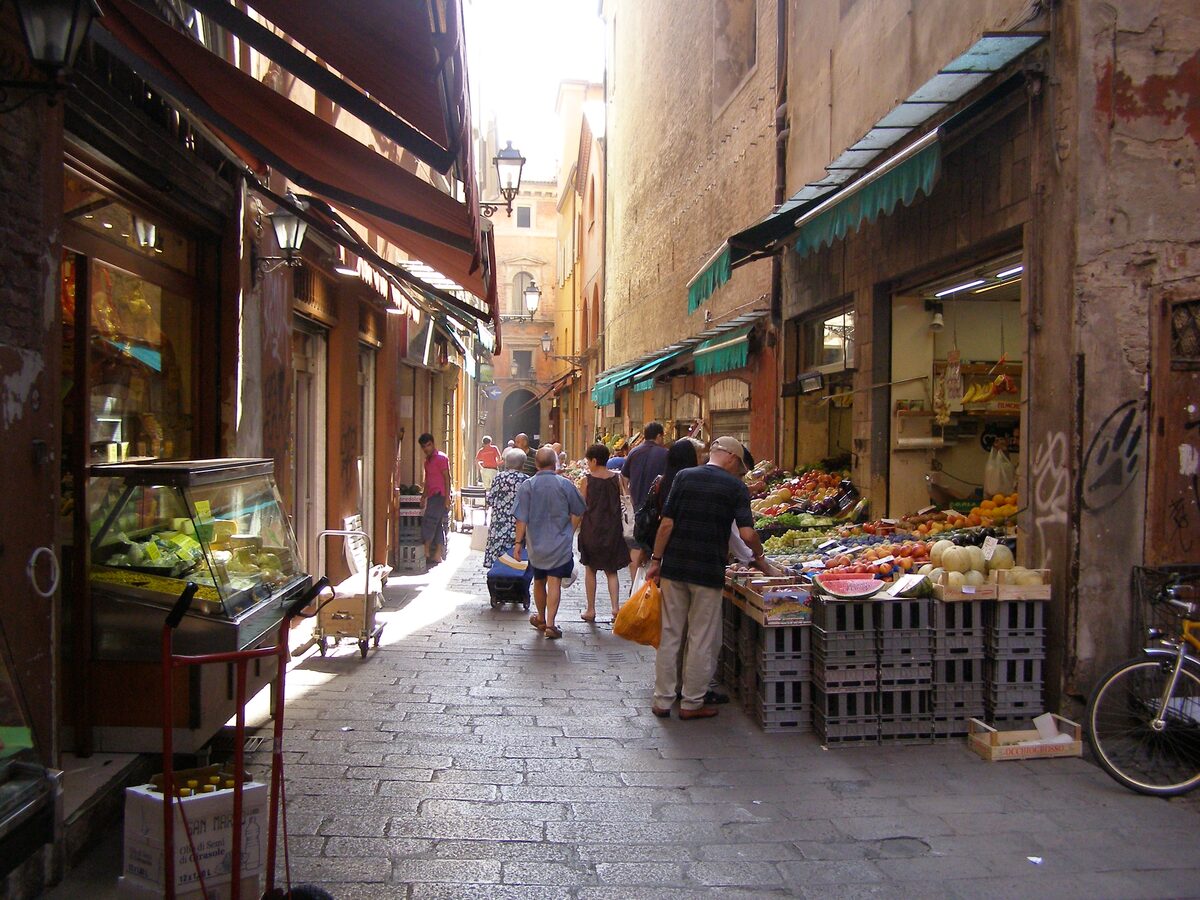 Shoppers walking a narrow market lane lined with fruit and vegetable stalls under awnings
