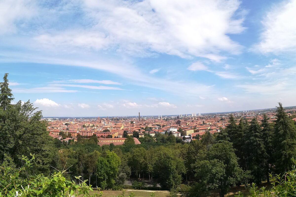 Bologna's red roofs and the Asinelli tower under a blue sky, seen over the trees from the hill of San Michele in Bosco