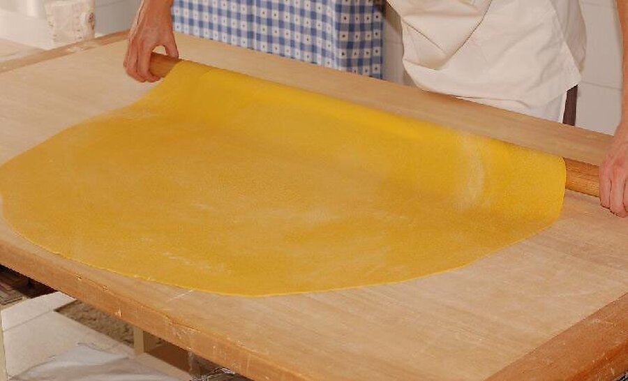 A cook rolling out a wide sheet of yellow egg pasta dough on a wooden board with a long rolling pin