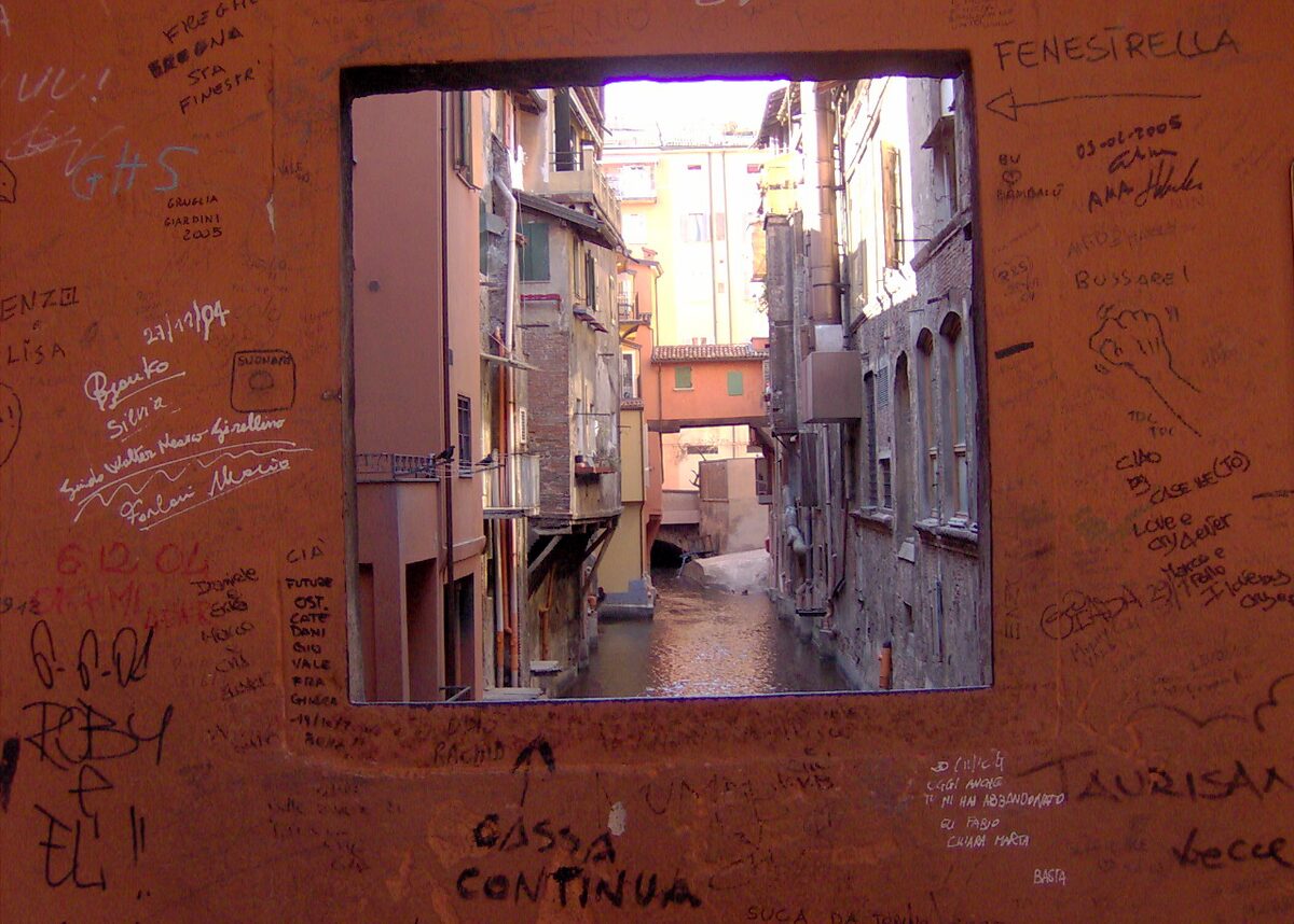 A small window cut into a graffiti-covered wall, framing a narrow street of old houses