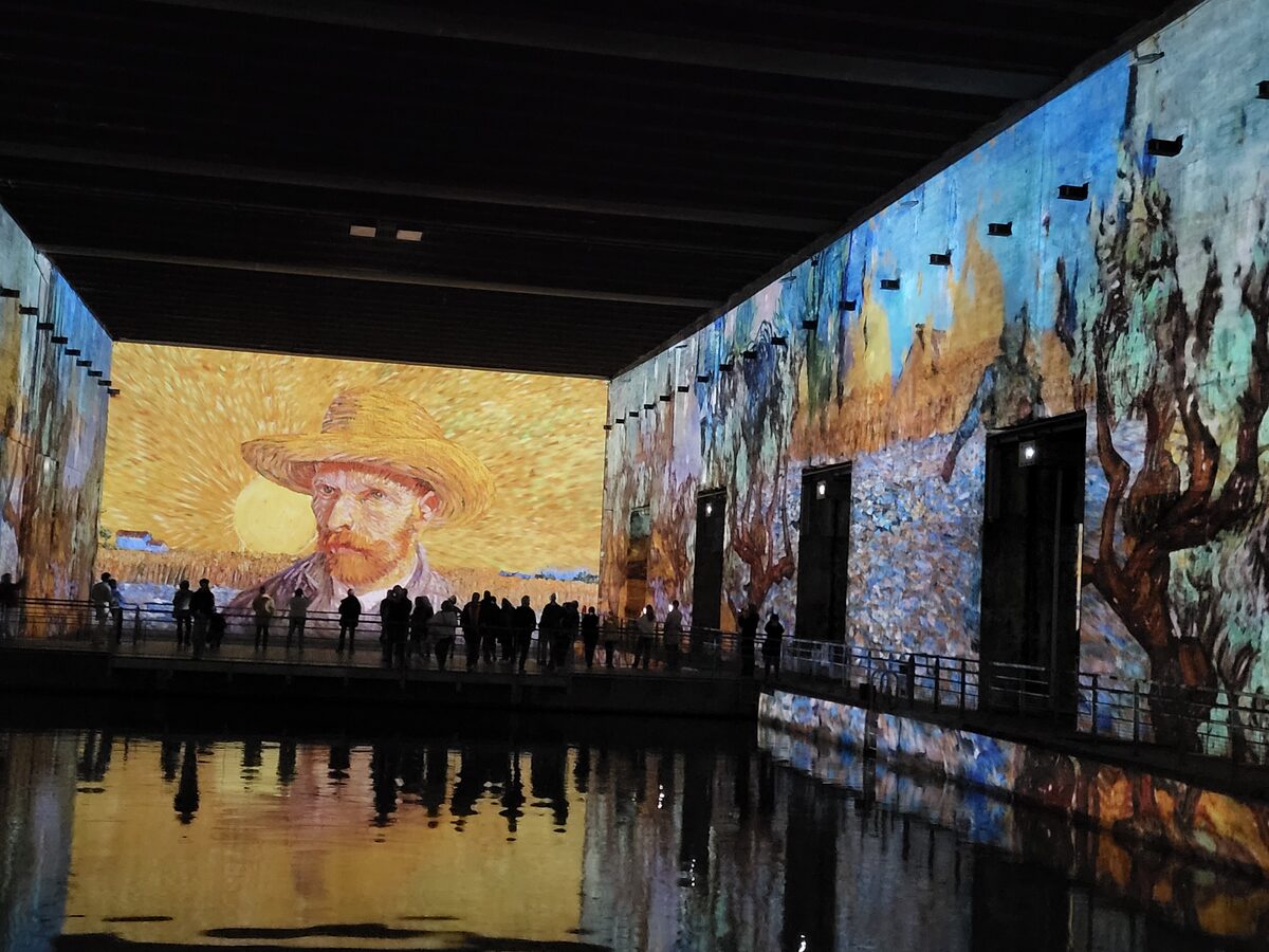 A Van Gogh self-portrait and wheat fields projected onto the walls of a vast dark concrete hall, reflected in the flooded basin below