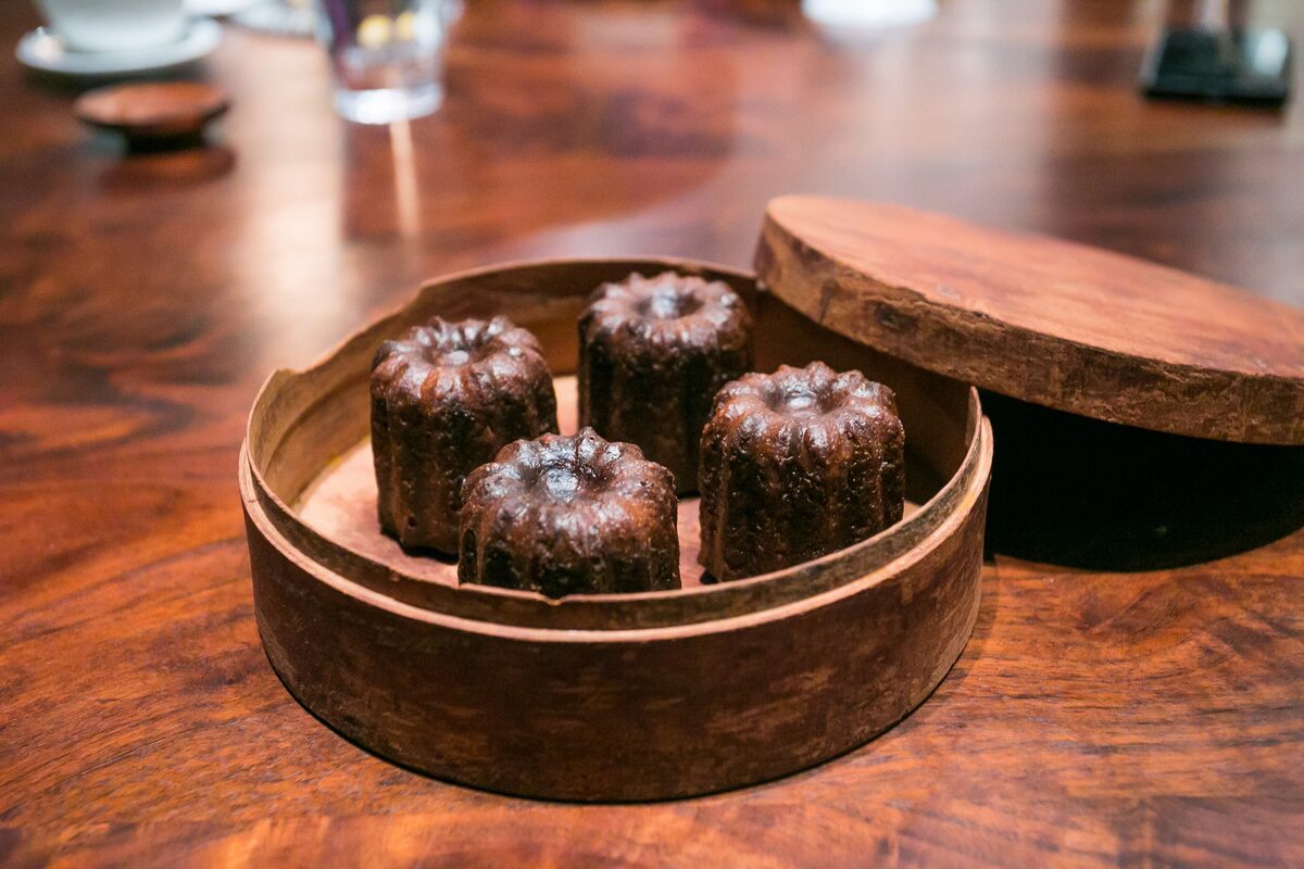 Three dark caramelized canelés in a round wooden box on a café table