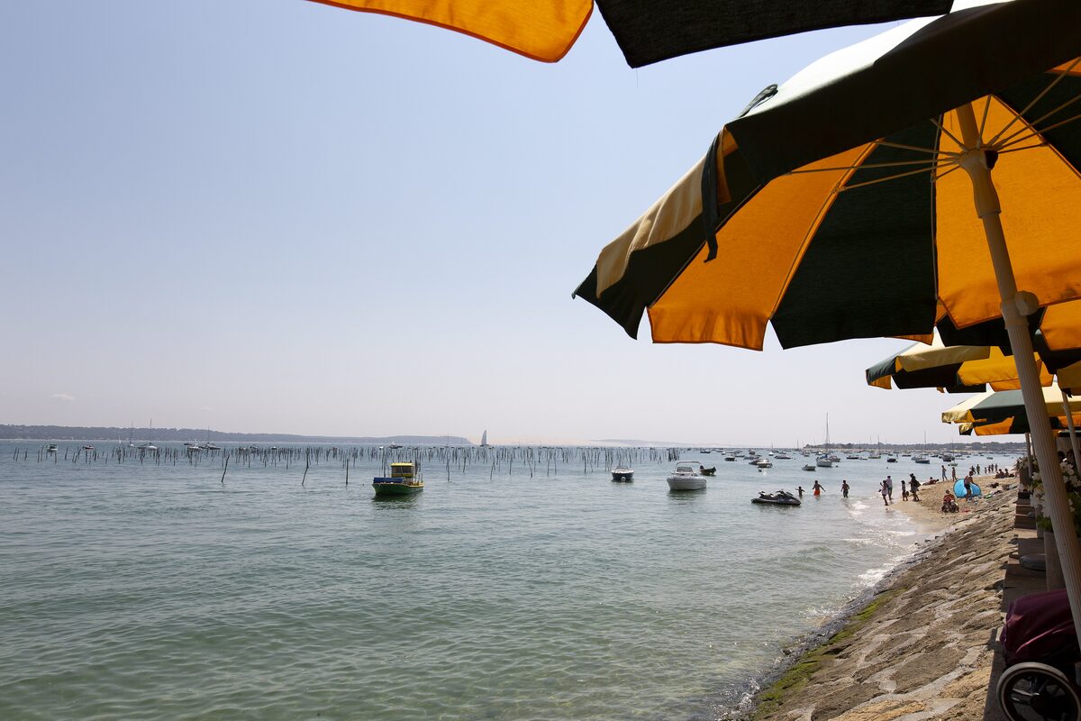 Arcachon Bay seen from a beach on Cap Ferret, with striped beach umbrellas, small boats and oyster stakes in the calm water