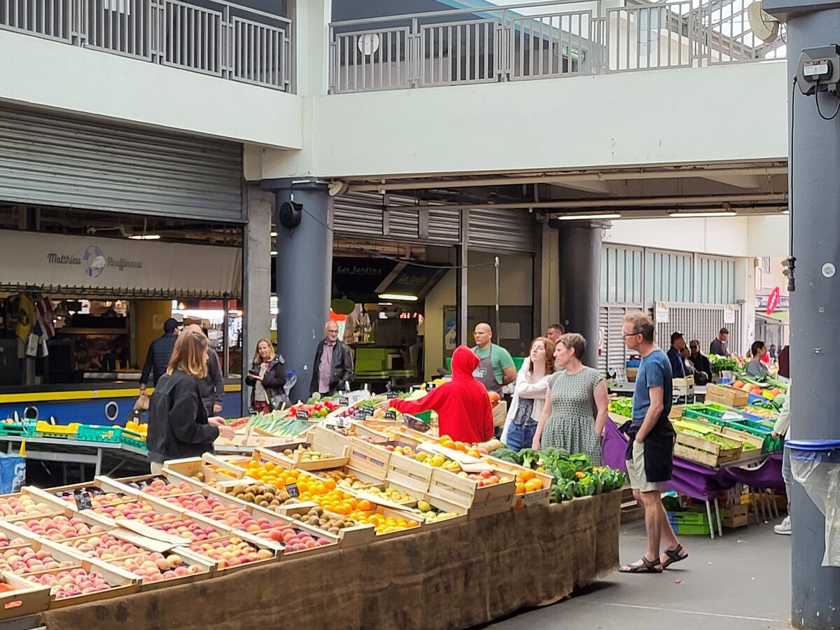 Crates of fruit and vegetables and shoppers at a produce stall inside the Marché des Capucins market hall