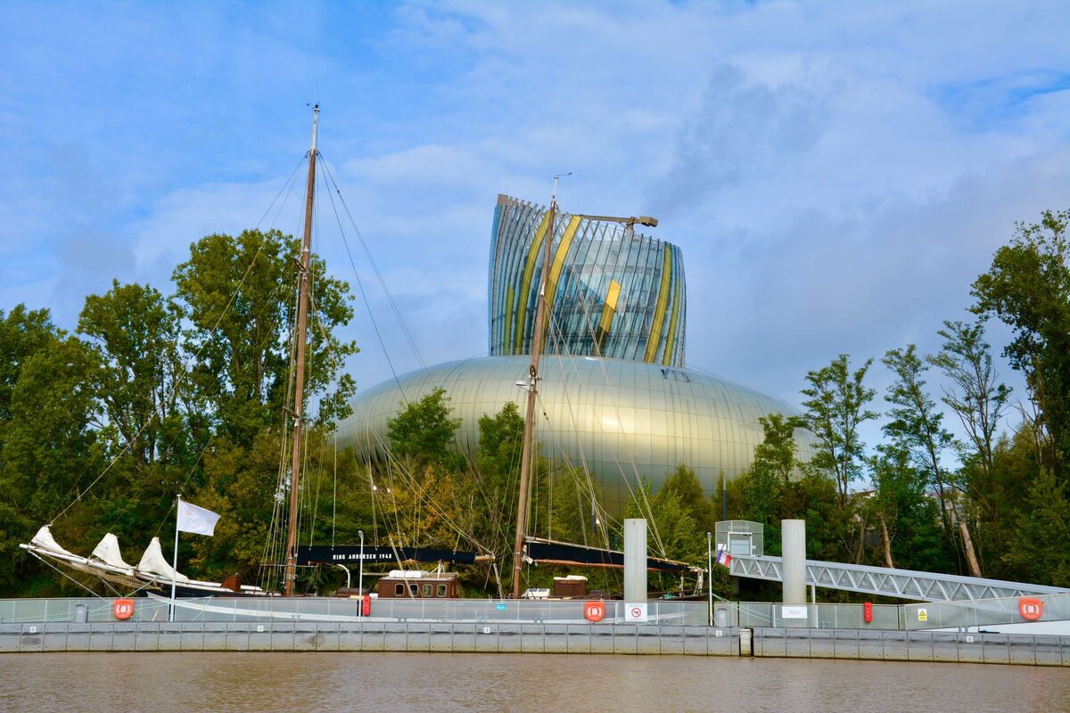 The curved gold and glass tower of La Cité du Vin rising above trees on the Garonne, with an old sailing boat moored in front