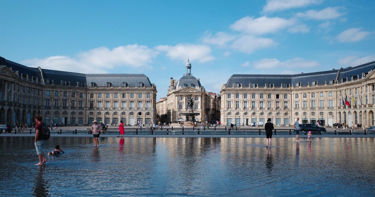 The Place de la Bourse in Bordeaux reflected in the shallow water of the Miroir d'eau, with people paddling in the water under a blue sky