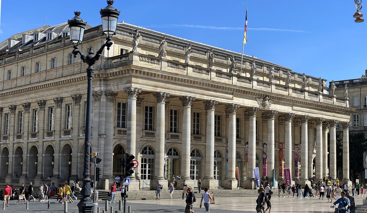 The neoclassical facade of the Grand Théâtre de Bordeaux with its twelve columns and statues on the roof, under a blue sky