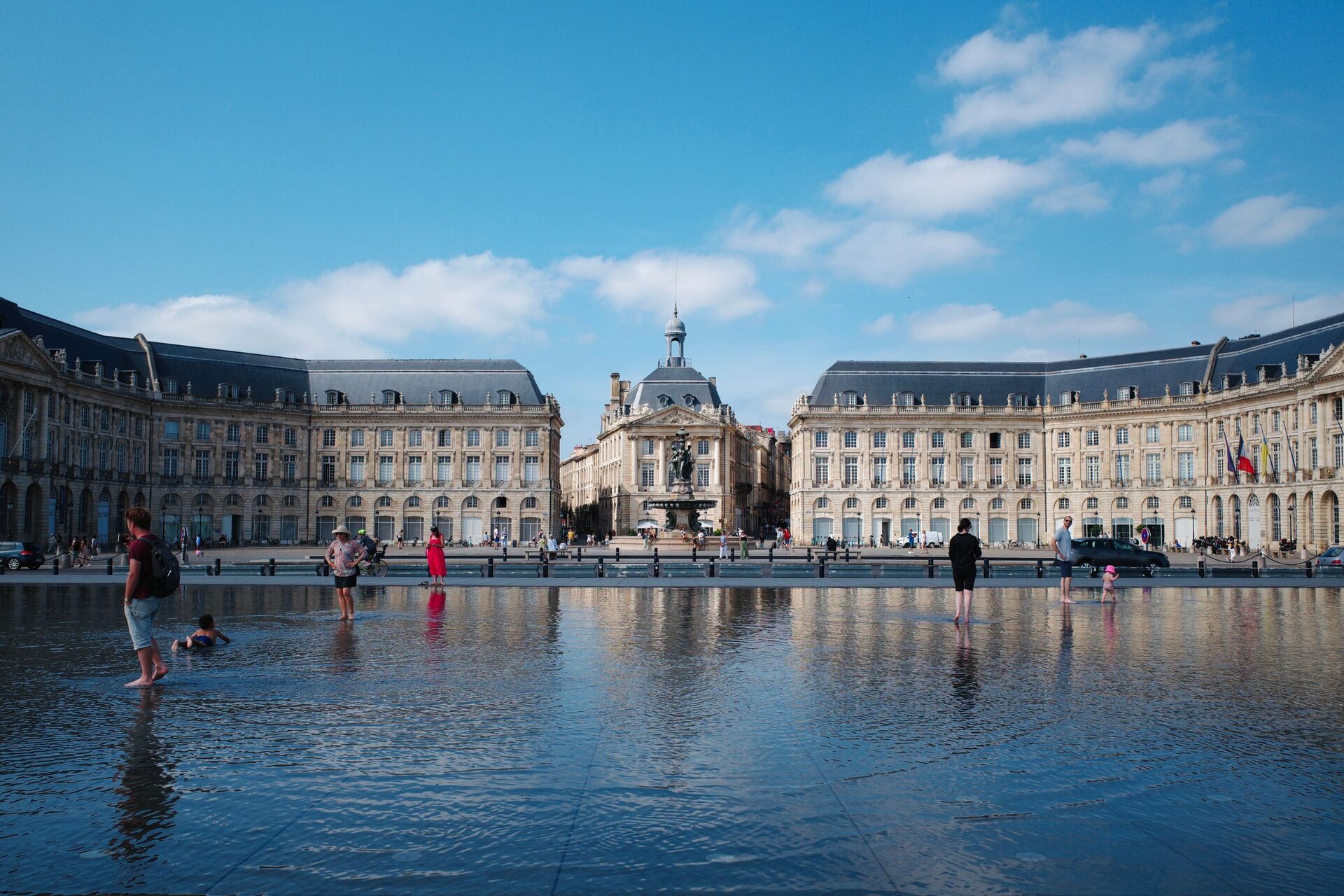 The Place de la Bourse in Bordeaux reflected in the shallow water of the Miroir d'eau, with people paddling in the water under a blue sky