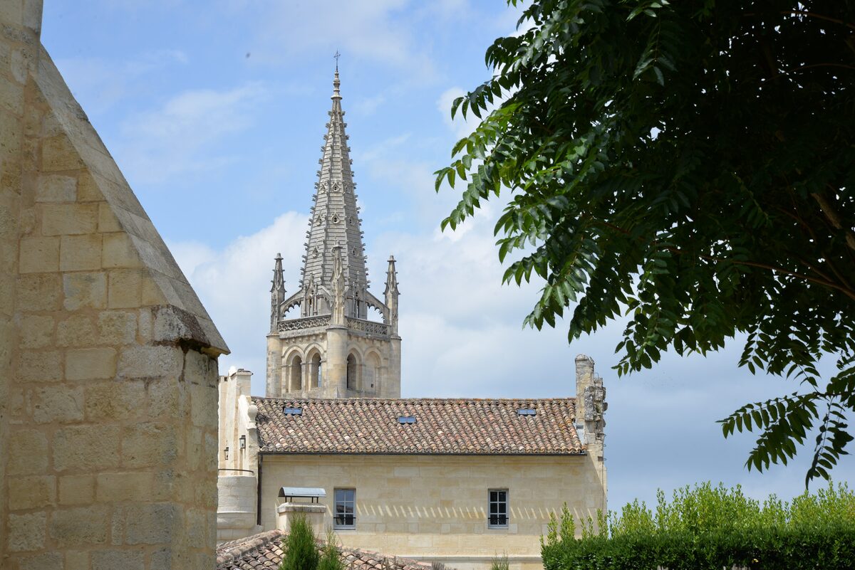 The tall Gothic spire of Saint-Émilion's monolithic church above a stone house and a tree, against a blue sky
