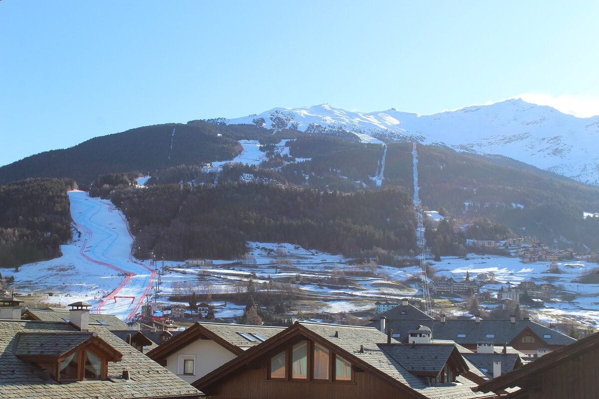 The red-netted Stelvio slope dropping from the wooded mountainside to the edge of Bormio, seen over chalet roofs with snowy peaks above