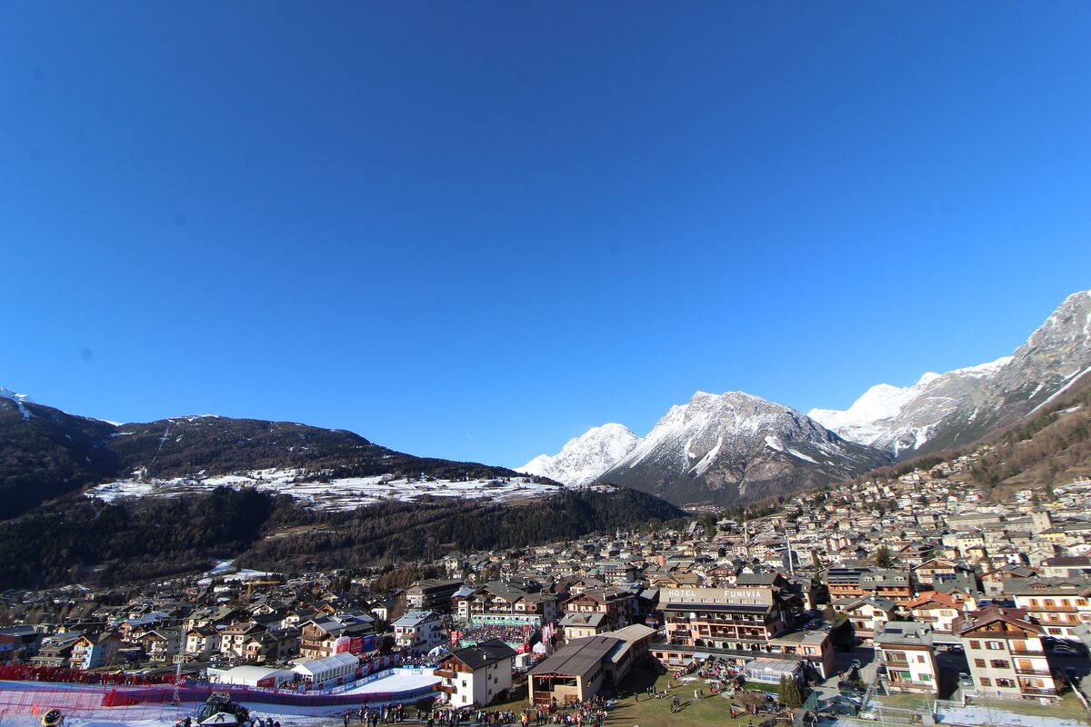 Bormio in December sunshine, seen from the hills above, with the town spread across the valley floor, a World Cup finish area on the left and a light cover of snow