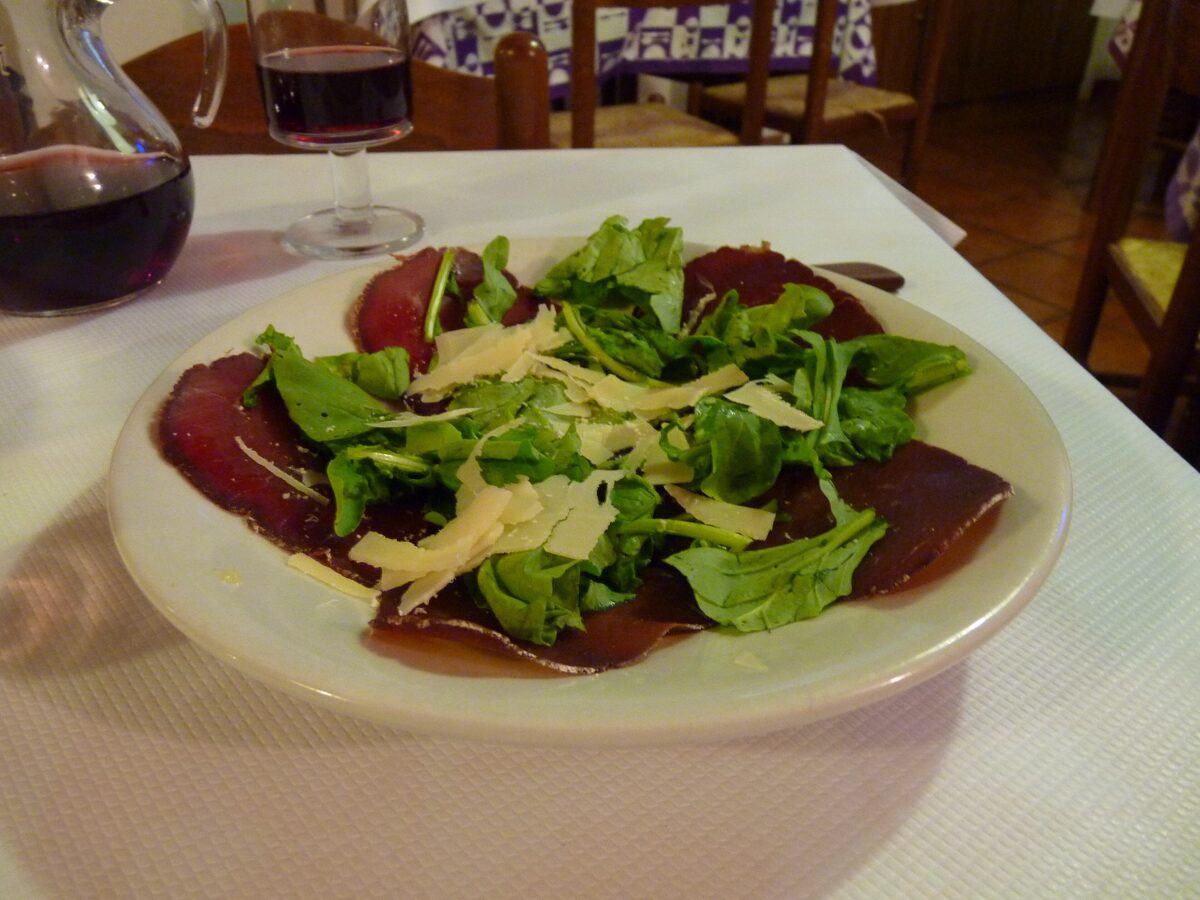 Thin slices of bresaola on a white plate with rocket and shaved Parmesan, with a glass of red wine behind