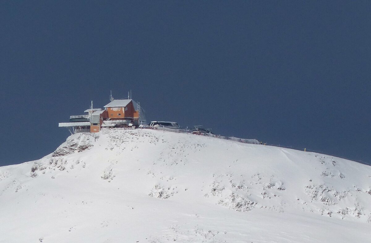 The top cable-car station of Bormio 3000 on a snowy summit, with an orange-brown building and a dark blue sky behind it, seen from a distance
