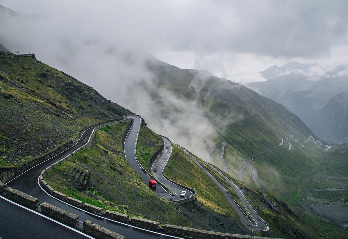 The hairpin bends of the Stelvio Pass road climbing through cloud and grassy slopes in summer; the pass is closed to cars in winter