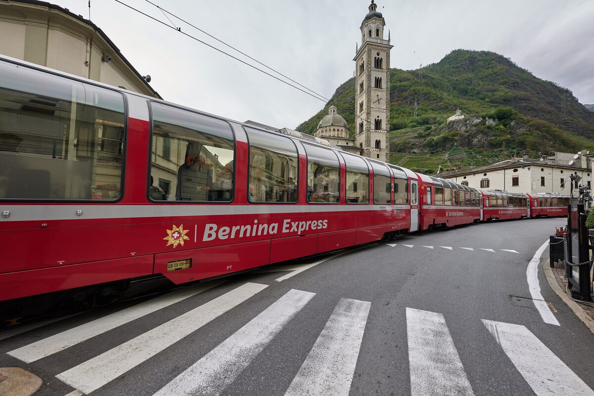 A red Bernina Express train running along the street in Tirano past the pilgrimage church and its tall bell tower