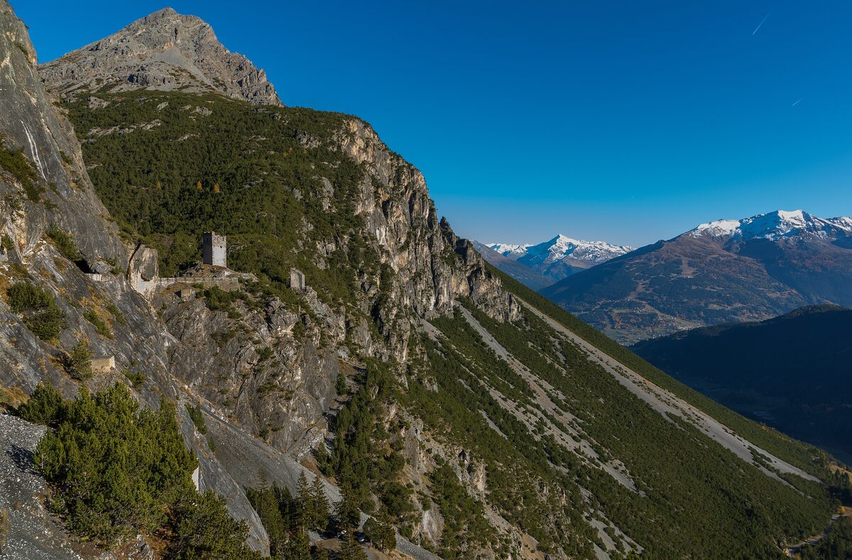 The north Fraele tower on a ridge above the road, with a sheer limestone cliff on the left and snow-dusted peaks in the distance