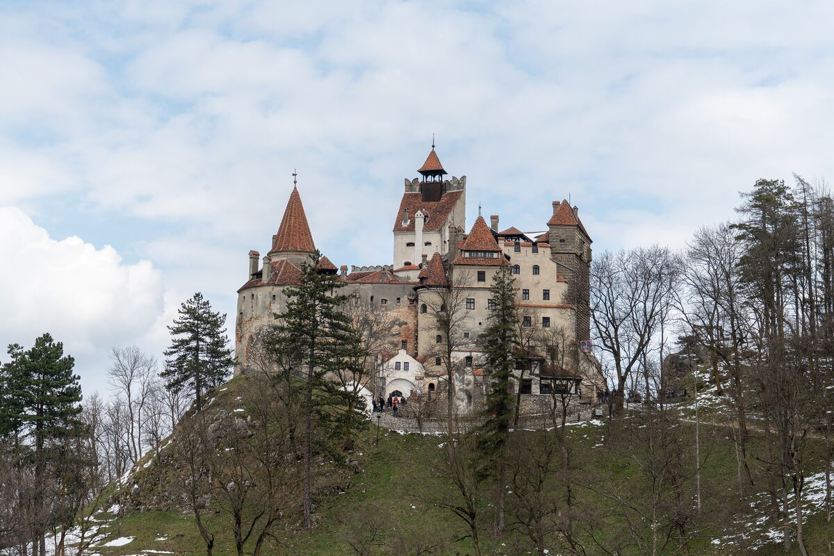 Bran Castle with its towers and red roofs on a rocky hill above the trees, under a blue sky with clouds