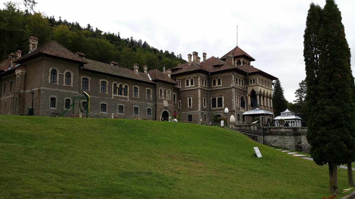 Cantacuzino Castle in Bușteni, a long stone mansion with red roofs and a tower, above a green lawn and dark fir trees