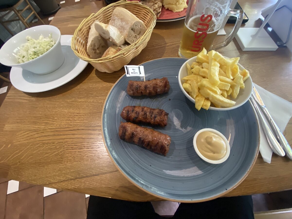 A plate of grilled mici with mustard, a bowl of fries and a glass of beer on a wooden table